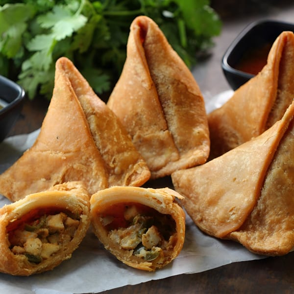 Golden-brown Indian samosas, one cut open to reveal a savory chicken and vegetable filling, with cilantro and sauce in the background.